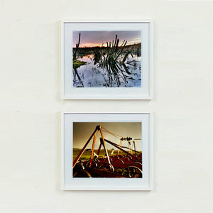 White framed photograph by Richard Heeps. Photograph of cut down, lichen clad branches poking out of the flooded fen field. The branches are strikingly dark and create dark reflections with a golden sunset in the background. The bottom photograph is of the wheels of an orangy/rusty looking plough bathed in the evening dusk light.