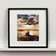 Load image into Gallery viewer, Black framed photograph by Richard Heeps. A tussock of grass sits at dusk in fenland water. It is siting under a black and white cloud formation with a golden dusk hue.