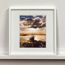 Load image into Gallery viewer, White framed photograph by Richard Heeps. A tussock of grass sits at dusk in fenland water. It is siting under a black and white cloud formation with a golden dusk hue.