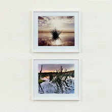 Load image into Gallery viewer, Two white framed photographs by Richard Heeps. The photograph on the top is of a tussock sitting in the water, black and reflected black into the fenland water below. The sky behind is dusky and atmospheric. The photograph at the bottom is of cut down winter sedge in a fenland waterway.