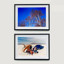 Load image into Gallery viewer, Two black framed photographs by Richard Heeps. The top photograph is looking up at the tops of four leafless silver birches against a deep blue autumn sky. The bottom photograph is a white desert with mountains in the back. At the centre of the photograph is a bright blue car with a small pod caravan behind and in front two women sitting in their camping chairs.