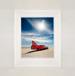 White framed photograph by Richard Heeps. A red drag car with a 75 written on its fin sits on a salt plain the front facing away towards the right. A blue cloudy sky is overhead.