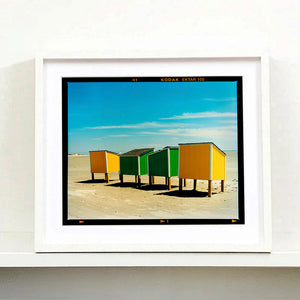 White framed photograph by Richard Heeps. Yellow and green beach lockers stand on their wooden legs on the clear sandy beach.