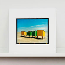 Load image into Gallery viewer, Mounted photograph by Richard Heeps. Yellow and green beach lockers stand on their wooden legs on the clear sandy beach.