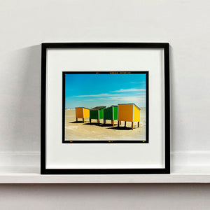 Black framed photograph by Richard Heeps. Yellow and green beach lockers stand on their wooden legs on the clear sandy beach.