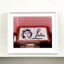 Load image into Gallery viewer, Black framed photograph by Richard Heeps. A kitsch Ladies&#39; toilet sign. The sign has the word Ladies alongside an outline of 1950s woman. The sign sits in a wooden frame against a pink tiled wall.