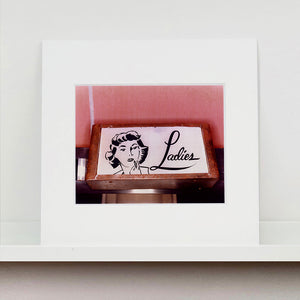 Mounted photograph by Richard Heeps. A kitsch Ladies' toilet sign. The sign has the word Ladies alongside an outline of 1950s woman. The sign sits in a wooden frame against a pink tiled wall.
