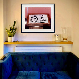 An in situ photograph by Richard Heeps. The photograph is situated on the wall behind a sofa. The photograph is of a kitsch Ladies' toilet sign. The sign has the word Ladies alongside an outline of 1950s woman. The sign sits in a wooden frame against a pink tiled wall.