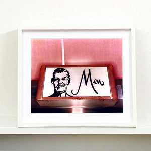 A white framed photograph by Richard Heeps. A kitsch Men's toilet sign. The sign has the word Men alongside an outline of 1950s man. The sign sits in a wooden frame and sits against a pink tiled wall.