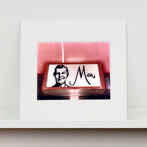 Mounted photograph by Richard Heeps. A kitsch Men's toilet sign. The sign has the word Men alongside an outline of 1950s man. The sign sits in a wooden frame and sits against a pink tiled wall.