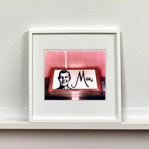 A white framed photograph by Richard Heeps. A kitsch Men's toilet sign. The sign has the word Men alongside an outline of 1950s man. The sign sits in a wooden frame and sits against a pink tiled wall.