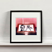 Load image into Gallery viewer, A black framed photograph by Richard Heeps. A kitsch Men&#39;s toilet sign. The sign has the word Men alongside an outline of 1950s man. The sign sits in a wooden frame and sits against a pink tiled wall.