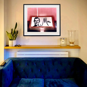 A black framed photograph by Richard Heeps is in situ behind a sofa. The photograph depicts a kitsch Men's toilet sign. The sign has the word Men alongside an outline of 1950s man. The sign sits in a wooden frame and sits against a pink tiled wall.