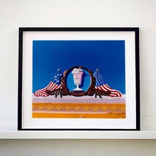 Load image into Gallery viewer, Black framed photograph by Richard Heeps. A 3D shape milkshake parlour sign which has a pink milkshake with a white top, cherry and straws, surrounded by a wooden type shield, and on either side the look of draped American flags. This is set against a blue sky.