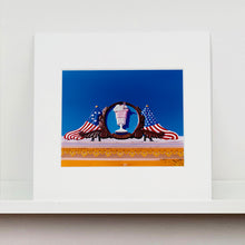 Load image into Gallery viewer, Mounted photograph by Richard Heeps. A 3D shape milkshake parlour sign which has a pink milkshake with a white top, cherry and straws, surrounded by a wooden type shield, and on either side the look of draped American flags. This is set against a blue sky.
