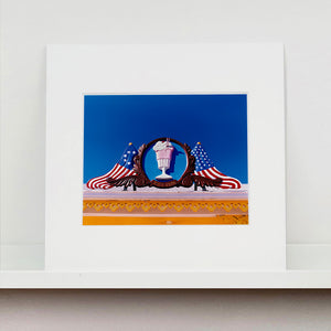 Mounted photograph by Richard Heeps. A 3D shape milkshake parlour sign which has a pink milkshake with a white top, cherry and straws, surrounded by a wooden type shield, and on either side the look of draped American flags. This is set against a blue sky.