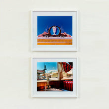Load image into Gallery viewer, Two white framed photographs by Richard Heeps. The top photograph is of a 3D shape milkshake parlour sign which has a pink milkshake with a white top, cherry and straws, surrounded by a wooden type shield, and on either side the look of draped American flags. This is set against a blue sky. The bottom photograph is the inside of a retro diner with the classic red seating. The view is looking out of the window onto an American small town road.