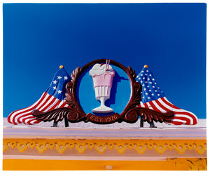 Photograph by Richard Heeps. A 3D shape milkshake parlour sign which has a pink milkshake with a white top, cherry and straws, surrounded by a wooden type shield, and on either side the look of draped American flags. This is set against a blue sky.