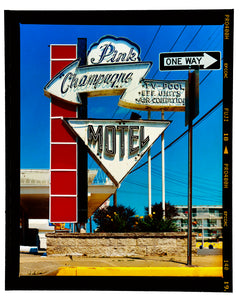 Photograph by Richard Heeps. A group of signs on an American Road sitting outside a motel mounted on a post with a vertical line of red squares.  The top of  the sign shows an arrow pointing to the left to Pink Champagne, there is an arrow pointing down with MOTEL written in it. In front of this motel sign is a one way sign pointing to the right.