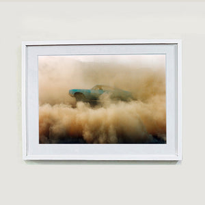 White framed photograph by Richard Heeps. A side view of a light blue Buick car moving and slightly obscured by the dust clouds which it has created.