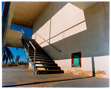 Load image into Gallery viewer, Photograph by Richard Heeps. The photograph has a metal staircase on the outside of a cream colour motel. The staircase has a ceiling but no sides so leads to blue sky. Behind the building is a blue sky and palm trees of the Californian Desert.