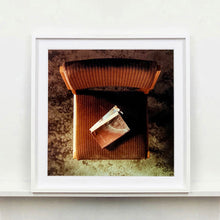 Load image into Gallery viewer, White framed photograph by Richard Heeps. The photograph looks down on a brown padded side chair. On it sits a waterstained and battered brown covered Holy Bible.