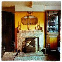Load image into Gallery viewer, Photograph by Richard Heeps. A vintage room with a fireplace with a brown tiled surround sitting in the middle of the photo flooded in daylight. On the mantelpiece stands 3 candlesticks, the walls behind are painted mustard yellow and there is a a decorative mirror. There is a dresser tucked into the recess on the right hand side.