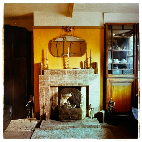 Photograph by Richard Heeps. A vintage room with a fireplace with a brown tiled surround sitting in the middle of the photo flooded in daylight. On the mantelpiece stands 3 candlesticks, the walls behind are painted mustard yellow and there is a a decorative mirror. There is a dresser tucked into the recess on the right hand side.