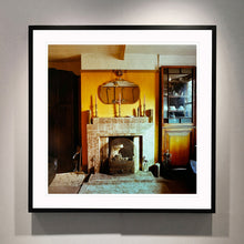 Load image into Gallery viewer, Black framed photograph by Richard Heeps. A vintage room with a fireplace with a brown tiled surround sitting in the middle of the photo flooded in daylight. On the mantelpiece sits 3 candlesticks, the walls behind are painted mustard yellow and there is a a decorative mirror. There is a dresser tucked into the recess on the right hand side.