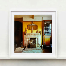 Load image into Gallery viewer, White framed photograph by Richard Heeps. A vintage room with a fireplace with a brown tiled surround sitting in the middle of the photo flooded in daylight. On the mantelpiece sits 3 candlesticks, the walls behind are painted mustard yellow and there is a a decorative mirror. There is a dresser tucked into the recess on the right hand side.