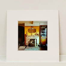 Load image into Gallery viewer, Mounted photograph by Richard Heeps. A vintage room with a fireplace with a brown tiled surround sitting in the middle of the photo flooded in daylight. On the mantelpiece sits 3 candlesticks, the walls behind are painted mustard yellow and there is a a decorative mirror. There is a dresser tucked into the recess on the right hand side.
