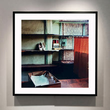 Load image into Gallery viewer, Black framed photograph by Richard Heeps. The photograph is of an abandoned shoe shop. Four rows of empty shoes shelves curve around a wall, apart from the middle shelf which has two mismatched white stilettos, and a small pile of paraphernalia. The walls are painted light green, held by darker green strutts. Retro brown curtains still hang in the window towards the right of the photograph. 