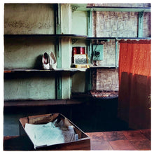 Load image into Gallery viewer, Photograph by Richard Heeps. The photograph is of an abandoned shoe shop. Four rows of empty shoes shelves curve around a wall, apart from the middle shelf which has two mismatched white stilettos, and a small pile of paraphernalia. The walls are painted light green, held by darker green strutts. Retro brown curtains still hang in the window towards the right of the photograph. 