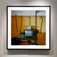Load image into Gallery viewer, Black framed photograph by Richard Heeps. Photograph of a vintage room, a television is on a table with two aerials on top, beside the television are drinks and glasses. Hanging from the picture rail on the yellow wall behind the television are 4 different walking sticks and a bed pan. 