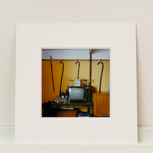 Load image into Gallery viewer, Mounted photograph by Richard Heeps. Photograph of a vintage room, a television is on a table with two aerials on top, beside the television are drinks and glasses. Hanging from the picture rail on the yellow wall behind the television are 4 different walking sticks and a bed pan. 