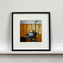 Load image into Gallery viewer, Black framed photograph by Richard Heeps. Photograph of a vintage room, a television is on a table with two aerials on top, beside the television are drinks and glasses. Hanging from the picture rail on the yellow wall behind the television are 4 different walking sticks and a bed pan. 