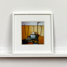 Load image into Gallery viewer, White framed photograph by Richard Heeps. Photograph of a vintage room, a television is on a table with two aerials on top, beside the television are drinks and glasses. Hanging from the picture rail on the yellow wall behind the television are 4 different walking sticks and a bed pan. 