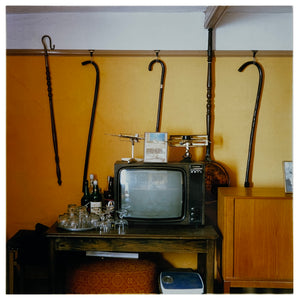 Photograph by Richard Heeps. Photograph of a vintage room, a television is on a table with two aerials on top, beside the television are drinks and glasses. Hanging from the picture rail on the yellow wall behind the television are 4 different walking sticks and a bed pan. 