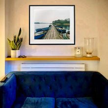 Load image into Gallery viewer, A framed Norfolk Broads photograph by Richard Heeps of a wooden dock extending over calm water with blue boats, displayed on a beige wall above a wooden shelf in a modern living room setting.