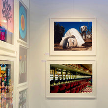 Load image into Gallery viewer, White framed photograph by Richard Heeps. In the foreground of the photograph is a broken off head of a giant fibreglass sculpture of a cowboy (a Muffler Man). In the background set in a deep blue sky is the muffler man&#39;s headless torso. The photograph is set amongst some other of Richard Heeps&#39; photographs exhibited at the Affordable Art fair.
