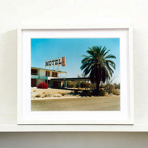 White framed photograph by Richard Heeps. A derelict motel office sits on a dusty American road. A large palm tree sits at the front of the office's walkway.