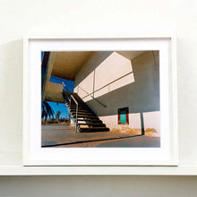 Load image into Gallery viewer, White framed photograph by Richard Heeps. The photograph has a metal staircase on the outside of a cream colour motel. The staircase has a ceiling but no sides so leads to blue sky. Behind the building is a blue sky and palm trees of the Californian Desert.