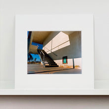 Load image into Gallery viewer, Mounted photograph by Richard Heeps. The photograph has a metal staircase on the outside of a cream colour motel. The staircase has a ceiling but no sides so leads to blue sky. Behind the building is a blue sky and palm trees of the Californian Desert.