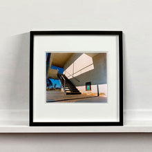 Load image into Gallery viewer, Black framed photograph by Richard Heeps. The photograph has a metal staircase on the outside of a cream colour motel. The staircase has a ceiling but no sides so leads to blue sky. Behind the building is a blue sky and palm trees of the Californian Desert.
