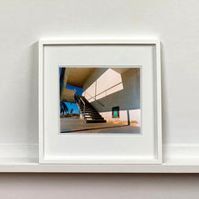 Load image into Gallery viewer, White framed photograph by Richard Heeps. The photograph has a metal staircase on the outside of a cream colour motel. The staircase has a ceiling but no sides so leads to blue sky. Behind the building is a blue sky and palm trees of the Californian Desert.