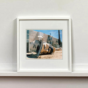 White framed photograph by Richard Heeps. The broken off head of a giant fibreglass sculpture of a cowboy (a Muffler Man) lies on the ground.