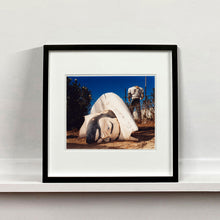 Load image into Gallery viewer, Black framed photograph by Richard Heeps. In the foreground of the photograph is a broken off head of a giant fibreglass sculpture of a cowboy (a Muffler Man). In the background set in a deep blue sky is the muffler man&#39;s headless torso.