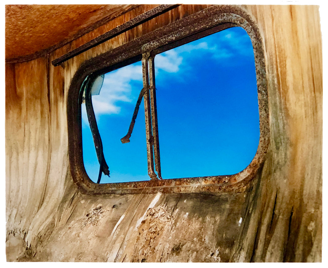 Photograph by Richard Heeps. The interior of an eroded trailer, looking through the broken window to a deep blue sky.