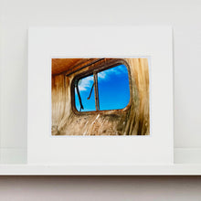 Load image into Gallery viewer, Mounted photograph by Richard Heeps. The interior of an eroded trailer, looking through the broken window to a deep blue sky.