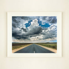 Load image into Gallery viewer, White framed photograph by Richard Heeps. A photograph looking along a long straight road into the distance, it is edged either side with sand and then prairie land. The vast, cloudy sky sits above.
